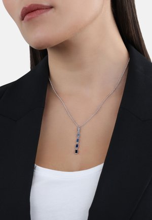 Collana pendente in argento con cinque pietre blu rettangolari, caratterizzata da una delicata catena e un design moderno e raffinato. Indossata sopra una maglietta bianca.