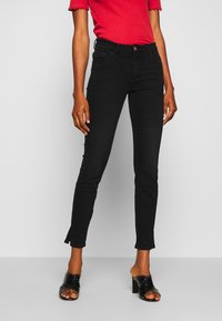 Jeans skinny de denim negro con cintura alta, que cuentan con un cierre de botón, un dobladillo deshilachado sutil, y se combinan con sandalias de tacón negro.