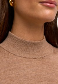 Maglione a dolcevita color cammello realizzato in tessuto a coste, con un colletto alto e una trama liscia. Primo piano del collo con delicati dettagli nelle cuciture.