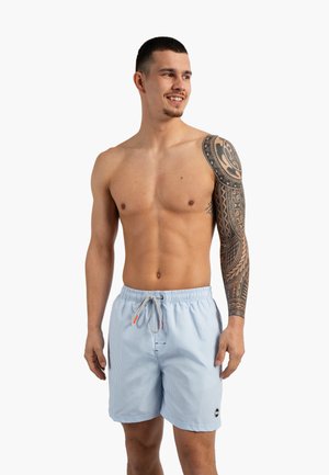 Jeune homme torse nu avec un tatouage tribal sur la manche porte un short de bain rayé bleu clair, souriant et regardant vers la droite sur fond blanc.