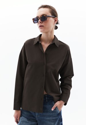 Bruine blouse met kraag, lange mouwen en een relaxed fit, gecombineerd met denim jeans en rechthoekige zonnebril.