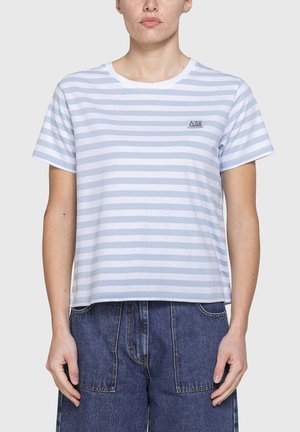Donna che indossa una maglietta a righe orizzontali bianche e azzurre e pantaloni di jeans blu scuro, in piedi con le mani rilassate lungo i fianchi.