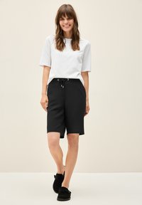 Svarta shorts med dragsko i midjan, ihop med en vit t-shirt med rund hals. Modellen bär svarta slip-on skor. Neutral bakgrund.