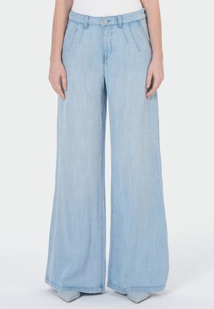 Lichtblauwe high-waist jeans met wijde pijpen, gedragen met puntige schoenen en een ingestoken wit topje, getoond van taille tot voeten.