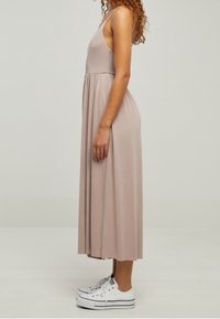 Femme debout de profil portant une robe midi sans manches mauve clair et des baskets compensées blanches sur un fond uni.