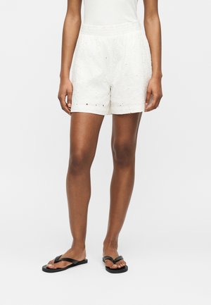 Femme portant un short blanc brodé, un haut blanc et des sandales plates noires, debout devant un fond uni.