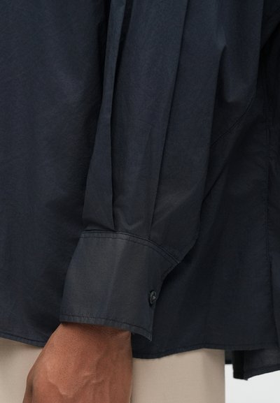 Tmavomodrá košeľa s dlhým rukávom, manžeta s gombíkom je zapnutá na zápästí osoby, pod lemom košele sú vidno béžové nohavice.