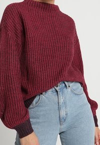 Maglione lavorato a maglia di un profondo bordeaux con un collo alto, texture a costine, maniche oversized e polsini scuri a contrasto, abbinato a jeans azzurri chiari.