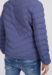 Doudoune bleu foncé avec un motif matelassé, présentant un col rond et un tissu texturé. Portée avec un jean bleu montrant des poches arrière.