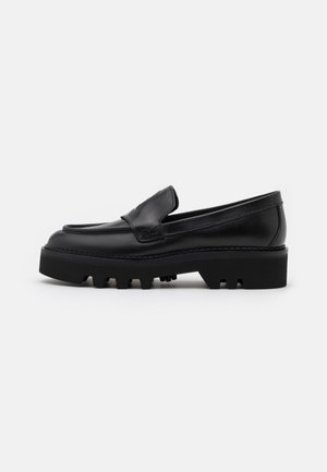 Mocassin en cuir noir avec semelle crantée épaisse et détail de coutures apparentes, présenté en profil latéral sur fond blanc.