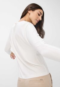 Lange mouwen, witte top van zachte stof. Heeft een relaxte pasvorm, achternaad details en een ronde halslijn. Gepaard met lichtbeige broeken.