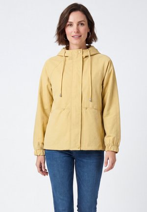 Frau mit kurzen braunen Haaren, die eine gelbe Kapuzenjacke mit Kordelzug und blaue Jeans trägt, steht und schaut nach vorne.
