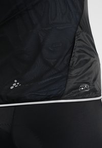 Gros plan d’une veste légère noire montrant un tissu en mesh, une bande réfléchissante et une icône de rangement pliable sur le bas du dos.