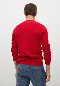 Roter Strickpullover mit gerippter Textur, rundem Ausschnitt und langen Ärmeln, kombiniert mit blauen Jeans. Keine sichtbaren Logos oder Muster.