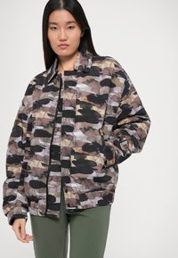 Jachetă matlasată cu un model de camuflaj multicolor în nuanțe de maron, negru și gri. Dispune de un fermoar frontal și manșete elastice.