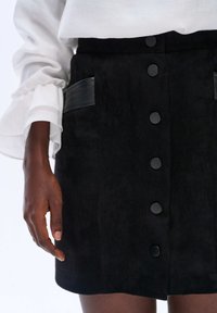 Zwart suedine mini-jurkje met een rechte snit, een sluiting met knopen aan de voorkant, twee zijzakken met zwarte leren accenten en een zachte textuur.