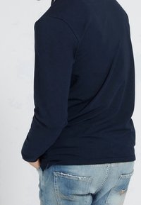 Man wearing navy long-sleeve shirt and light blue distressed jeans with hands in pockets, viewed from the back against plain background.