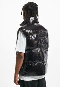 Gilet imbottito nero lucido con collo alto, caratterizzato da sezioni trapuntate e vestibilità ampia, indossato sopra una T-shirt bianca.