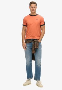 Orange bomulds t-shirt med marineblå stribet krave, parret med lyseblå slidte jeans og en ternet skjorte knyttet om taljen. Beige sneakers.