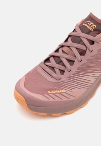 Light pink athletic shoe featuring a breathable mesh upper, textured accents, and a pronounced orange outsole with a rugged tread pattern.
