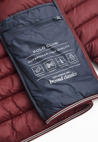 Bordeauxfärgad kviltad jacka med marinblå innerfoder som visar skötselråd och logotyp, accentuerad med en trefärgad rand.