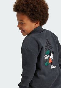 Giacca in fleece grigia scura con un design ricamato di Topolino e il logo Adidas sul retro. Texture morbida con polsini elasticizzati.