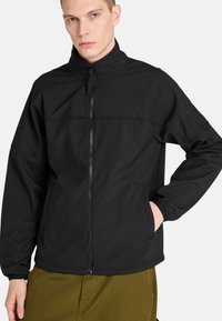 Musta pehmeäkuorinen takki korkealla kauluksella, etuvetoketjulla ja sivutaskuilla. Sisältää huomaamattomia ompeluyksityiskohtia ja rentoa istuvuutta.