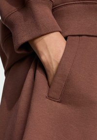 Hnedá mikina s rebrovanými manžetami, mäkkou textúrou a bočnou kapsou, pričom viditeľná ruka je čiastočne zasunutá do vrecka.