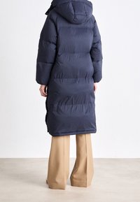 Marineblauer Steppmantel mit Kapuze, mit gestepptem Design und langen Ärmeln, der bis zur Mitte der Wade reicht. Kombiniert mit beigen weit geschnittenen Hosen.