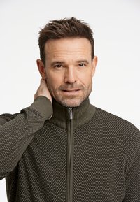 Grön zip-tröja med texturerat mönster, hög krage och ribbade ärmslut. Den har en dragkedja framtill och är tillverkad av ett mjukt material.