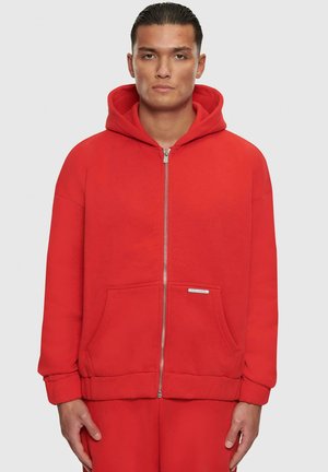 Rød zip-up hoodie lavet af blødt materiale, med en kængurulomme, en stor hætte og ribbede manchetter. Klar brandingmærke nær lommen.