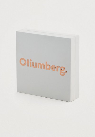 Γκρι κουτί με λείο φινίρισμα, με το όνομα της μάρκας "Otiumberg" σε ανάγλυφα γράμματα ροζ χρυσού στο μπροστινό μέρος. Τετράγωνο σχήμα.