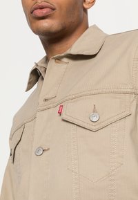 Veste en denim beige avec col à revers, boutons argentés, deux poches poitrine et étiquette de logo de marque rouge. Présente une texture côtelée verticale.