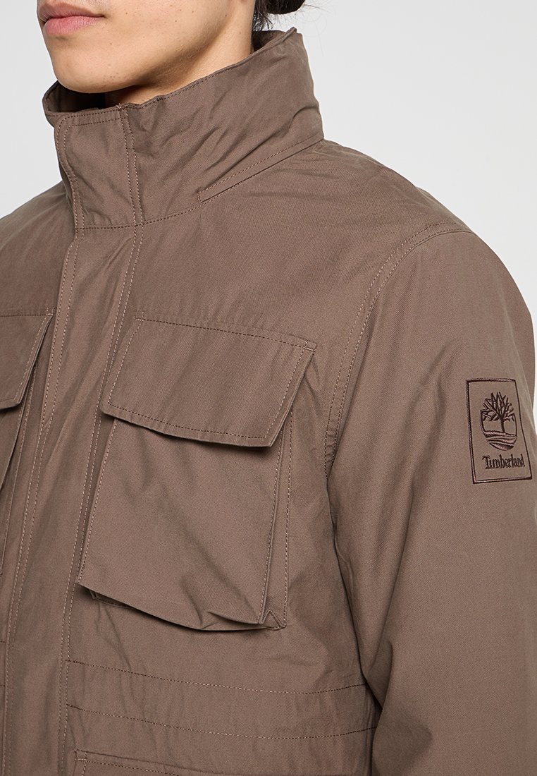 Giacca marrone, impermeabile, con colletto alto, due tasche frontali e patch logo ricamato sulla manica. Texture liscia, design strutturato.