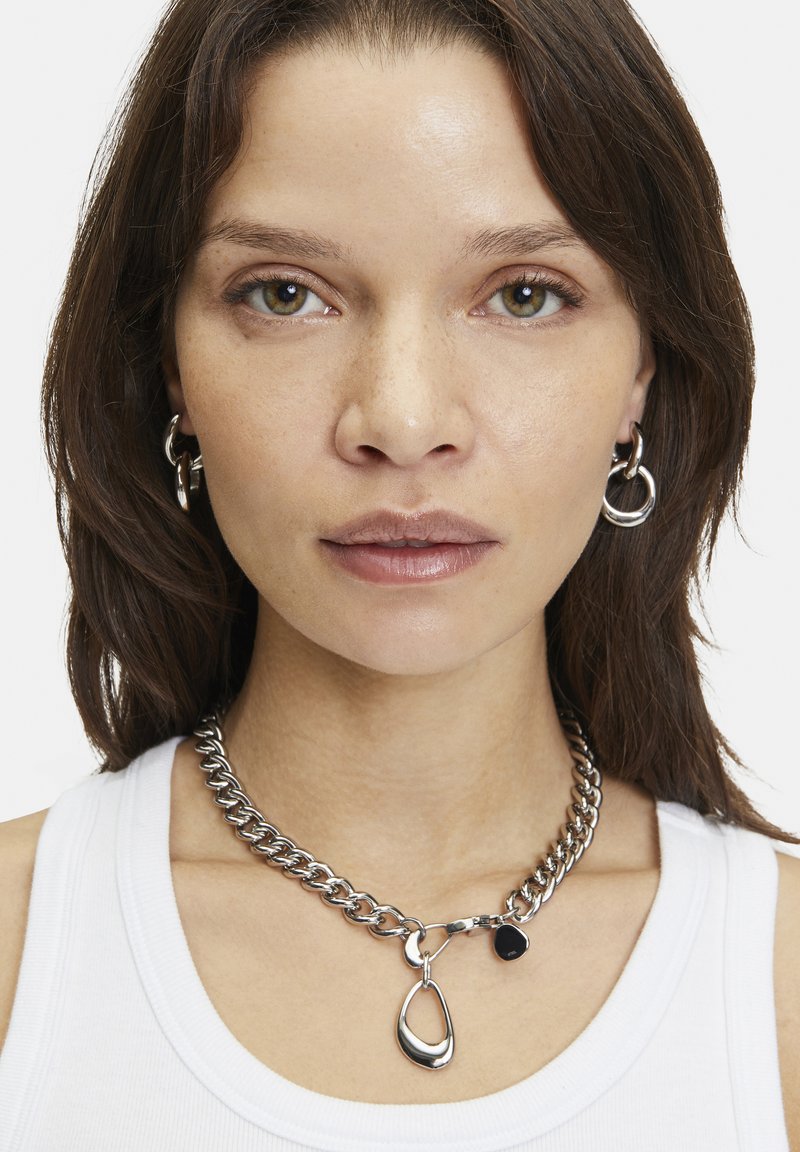 Sølvmetal halskæde med chunky kædeled og en dråbe vedhæng, parret med matchende hoops øreringe. Modellen bærer en hvid top.