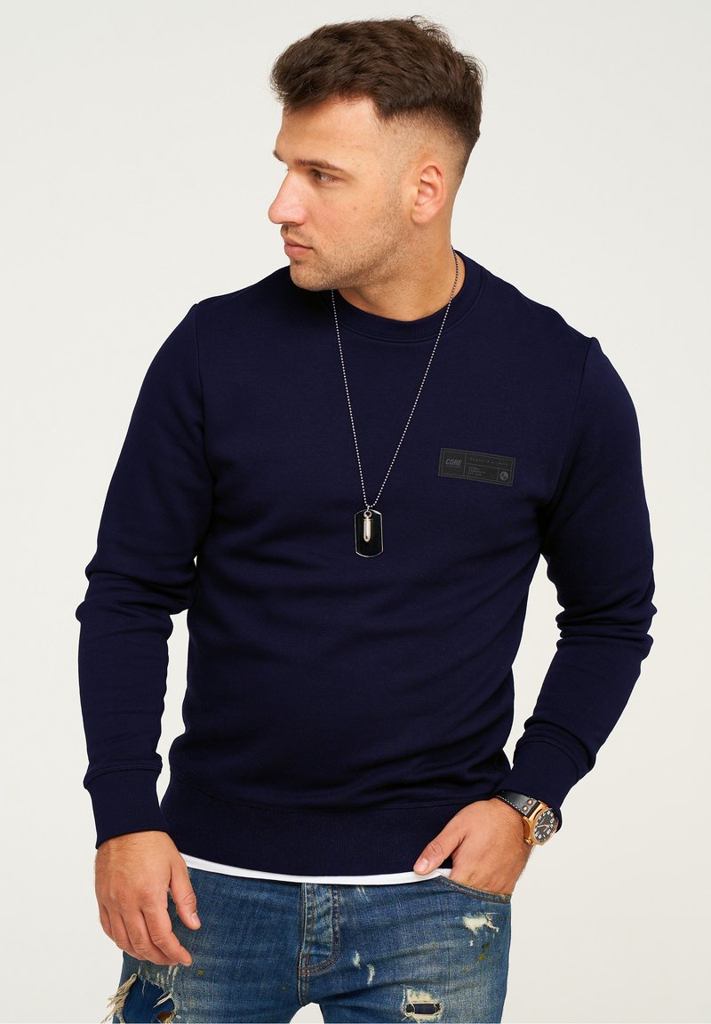 Navy blue sweatshirt, crew neck, ribbed cuffs and hem, features a black label on the chest. Worn with distressed denim and a watch.
