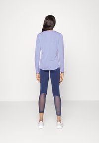 Lila Langarmoberteil kombiniert mit marineblauen Leggings, die transparente Mesh-Einsätze aufweisen. Getragen mit weißen Sportschuhen. Ansicht von hinten.