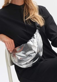 Femme portant une tenue noire assise sur une chaise, tenant un sac porté travers argenté brillant avec une bandoulière sur l'épaule.