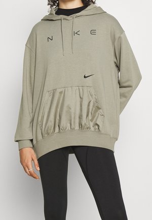 Personne portant un sweat à capuche Nike vert sauge avec poche avant et leggings noirs, main détendue sur le côté, torse visible sans visage.