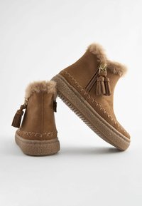 Stivaletti alla caviglia in suede marrone con rifiniture in pelliccia sintetica, dettaglio con nappine e suola in gomma testurizzata. Presentano una cerniera laterale per una facile vestibilità.