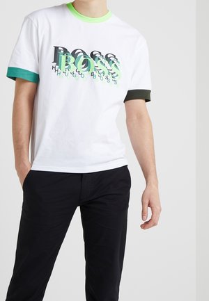Mann trägt weißes T-Shirt mit grünen und schwarzen Ärmelsäumen und mehrfarbigem "BOSS"-Schriftzug, kombiniert mit schwarzen Hosen, steht vor einfarbigem Hintergrund.