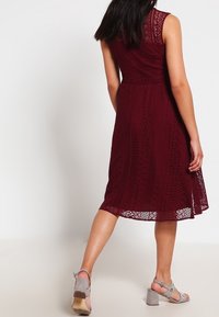 Burgunderfärgad spetsklänning med ärmlös design, som har en figurnära midja och en flödande kjol. Utsmyckad med blommönster och scalloped fåll.