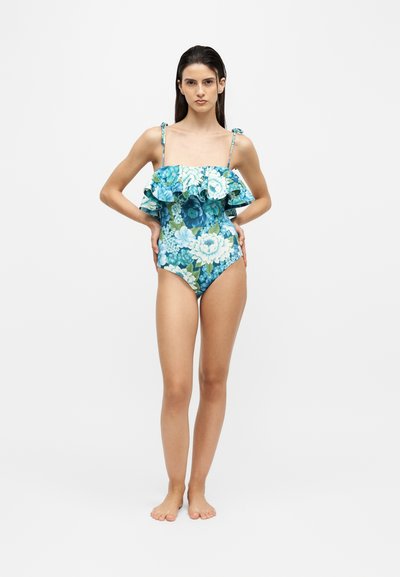 Femme debout pieds nus portant un maillot de bain une pièce bleu à motif floral avec haut à volants et bretelles réglables sur fond blanc.