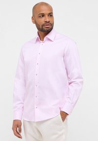 Helles rosa langärmliges Hemd mit vertikalen weißen Streifen, Button-down-Kragen und silbernen Knöpfen entlang der Vorderseite, kombiniert mit cremefarbenen Hosen.