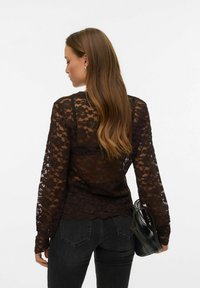 Črna čipkasta majica z dolgimi rokavi, z nazobčanim robom in detajli, združena s temnimi kavbojkami in črno torbico. Vidna je teksturirana cvetlična vzorec.