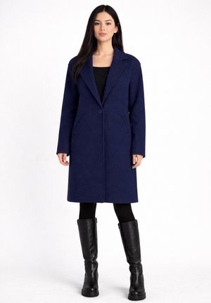 Donna in piedi che indossa un cappotto blu navy fino al ginocchio, leggings neri e stivali neri alti fino al ginocchio su uno sfondo bianco semplice.
