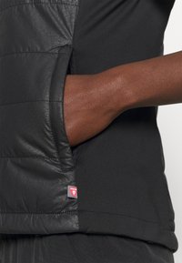 Svart jacka med vadderat textil på ena sidan och ett mjukt, stretchigt tyg på den andra sidan. Inkluderar en sidficka. Röd logotypetikett synlig.