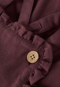 Tessuto bordeaux con una morbida texture, caratterizzato da dettagli arricciati e un bottone di legno con quattro fori, che mette in risalto la sua particolare cucitura.