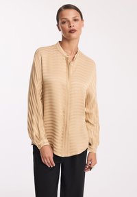 Beżowa bluzka z długim rękawem z delikatnym prążkowanym wzorem, z zapięciem na guziki i kołnierzykiem mandaryńskim, zestawiona z czarnymi spodniami.