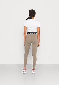 Beige legging met een hoge zwarte tailleband met het label "NIKE," gedragen met witte sportschoenen met roze accenten. Effen wit T-shirt.